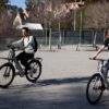 Un hombre y una mujer montando en bicicletas eléctricas modernas por un camino pavimentado en un entorno urbano con edificios modernos de fondo.