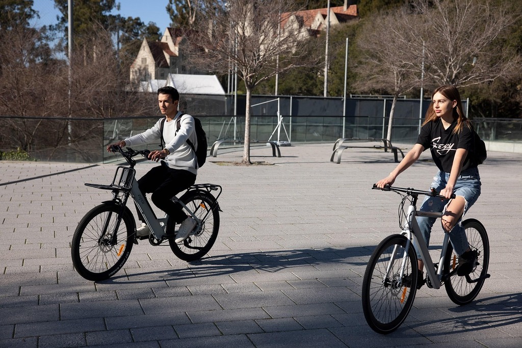 Un hombre y una mujer montando en bicicletas eléctricas modernas por un camino pavimentado en un entorno urbano con edificios modernos de fondo.