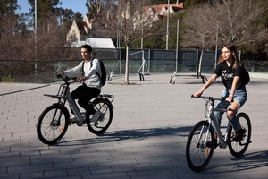 Un hombre y una mujer montando en bicicletas eléctricas modernas por un camino pavimentado en un entorno urbano con edificios modernos de fondo.