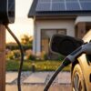 Primer plano de un coche eléctrico cargándose en un cargador de pared doméstico con una casa con paneles solares al fondo al atardecer