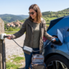 Fotografía de una mujer sonriente cargando un coche eléctrico azul en una zona de descanso de montaña con vistas panorámicas y un pueblo rural al fondo.