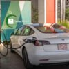 Un Tesla Model 3 blanco está estacionado y conectado a un cable de carga en una gran estación de carga de vehículos eléctricos PuntoElectric, con un letrero verde y azul al fondo.