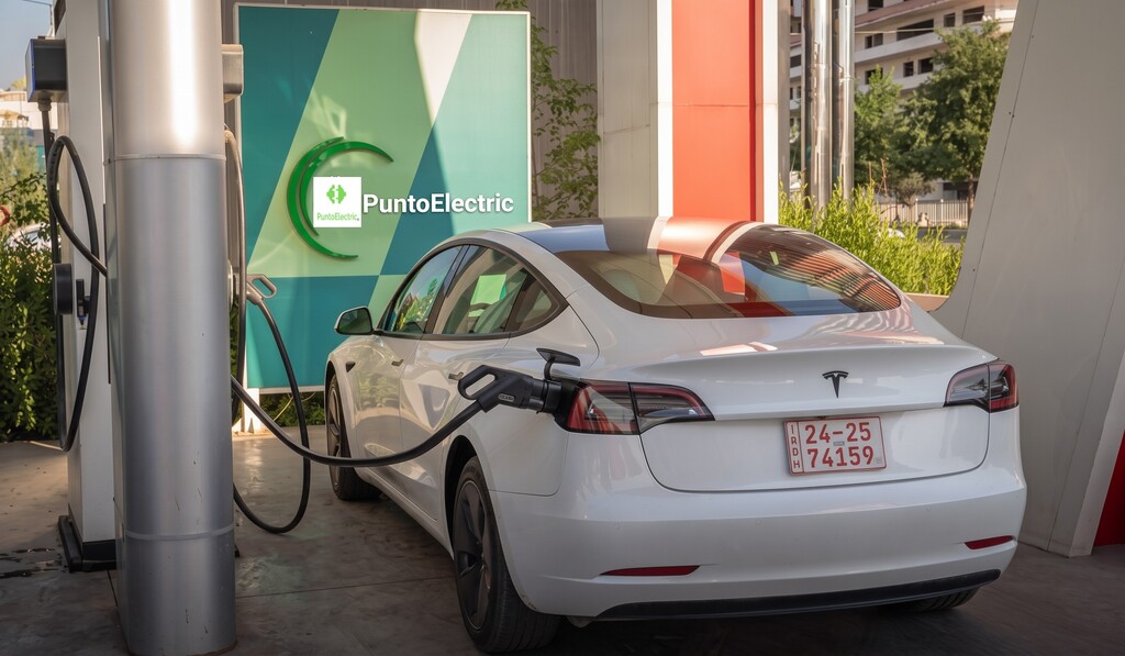 Un Tesla Model 3 blanco está estacionado y conectado a un cable de carga en una gran estación de carga de vehículos eléctricos PuntoElectric, con un letrero verde y azul al fondo.