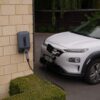 Coche eléctrico blanco conectado a un punto de recarga de pared en el exterior de una vivienda.