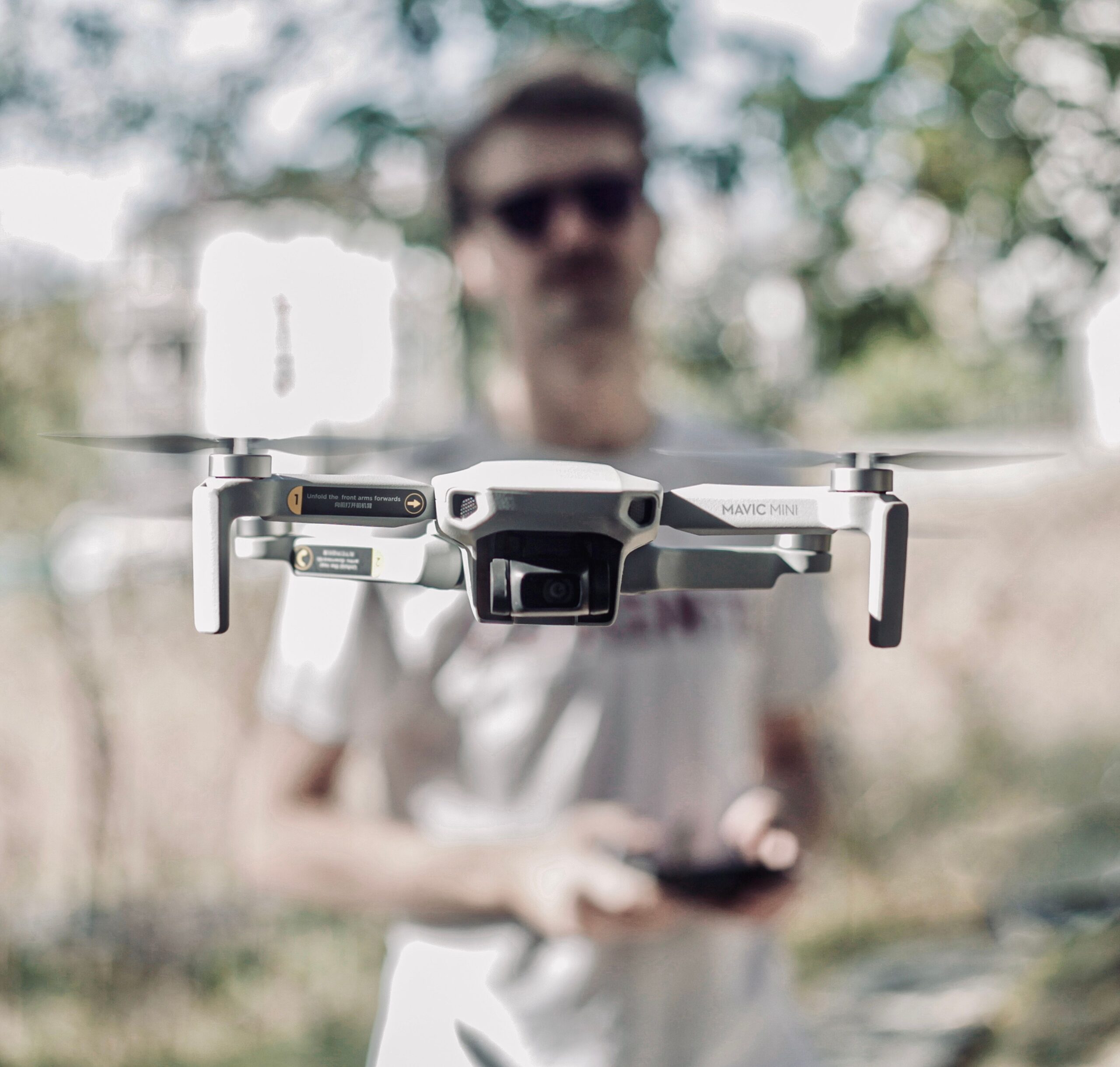 Primer plano de un dron ligero en vuelo mientras un piloto lo maneja al fondo al aire libre.